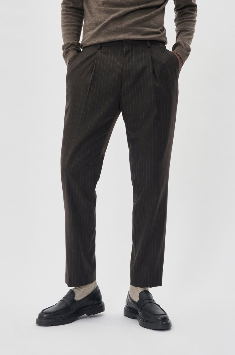 Braune Nadelstreifenhose mit taillierter Passform, versehen mit Falten an der Vorderseite. Kombiniert mit dunklen Slippern und hellen Socken.