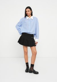 Svetlomodrá pruhovaná košeľa na gombíky s bielym golierom, skombinovaná s čiernou plisovanou sukňou a lesklými čiernymi topánkami.