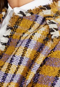 Chal tejido en patrón de cuadros en amarillo, púrpura y negro con bordes deshilachados. La tela tiene un acabado texturizado con hilos multicolores.