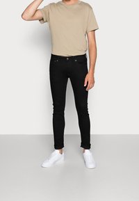 Muž v béžovém tričku, černých slim-fit džínách na ohrnutých koncích a bílých teniskách, stojící s jednou rukou zvednutou k hlavě.