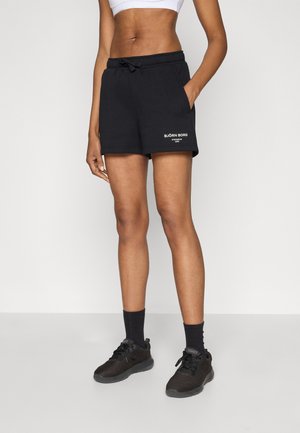 Mustad dressipüksid, millel on elastne vöökoht ja nöör, küljetaskud ja vasaku jala valge tekstibränding.