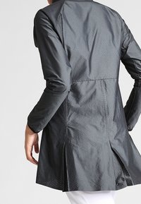 Chaqueta negra ligera con una textura suave, que presenta un diseño acampanado, perforaciones discretas y aberturas laterales para facilitar el movimiento.