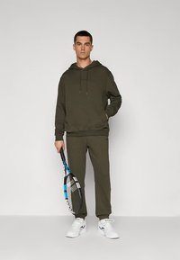 Zielona oliwkowa bluza z kapturem z troczkami i przednią kieszenią, zestawiona z pasującymi spodniami dresowymi. Białe buty sportowe i rakieta tenisowa dopełniają stylizację.