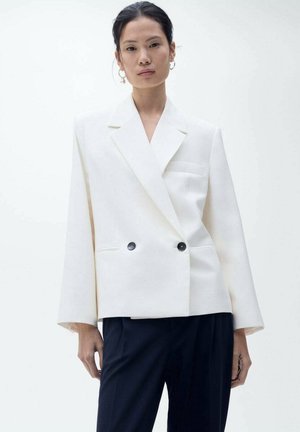 Double-breasted white blazer with notched lapels, two black buttons, and long sleeves, paired with dark trousers. Smooth fabric texture.