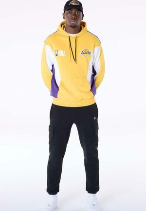 Uomo che indossa una felpa con cappuccio gialla e viola dei Los Angeles Lakers, pantaloni cargo neri, sneakers bianche e un berretto nero dei Lakers, in piedi con le mani dietro la schiena.