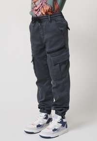 Temno sivi cargo hlače z elastičnimi manšetami, ki imajo več žepov in zadrgo. Kombinirane z belimi in mornarsko modrimi visokimi supergami.