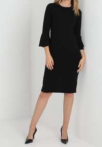 Černé šaty ke kolenům s dlouhými, rozšířenými rukávy, kulatým výstřihem a přiléhavým střihem. Nosené s černými vysokými podpatky.