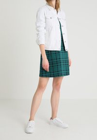 Femme portant une veste en denim blanche, une robe courte à carreaux verts et des baskets blanches, se tenant devant un fond clair.