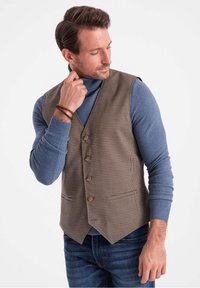 Braunes Hahnentrittmuster-Westen mit vier Knöpfen, getragen über einem blauen Rollkragenpullover. Die Weste hat eine taillierte Passform und zwei Fronttaschen.