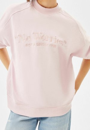 Person iført en lyserød sweatshirt med teksten "No Worries" og "Not a Single One" broderet foran, kombineret med lyseblå jeans.