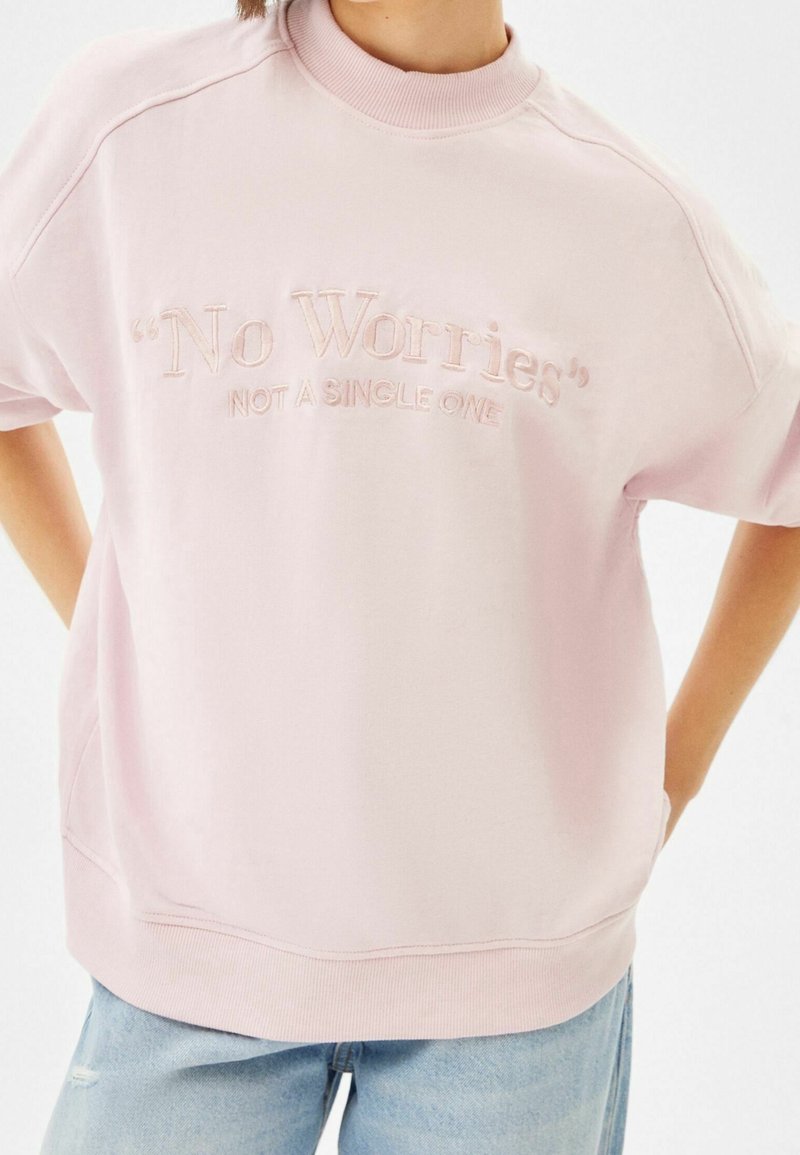 Personne portant un sweat-shirt rose clair avec les textes "No Worries" et "Not a Single One" brodés sur le devant, associé à un jean bleu clair.