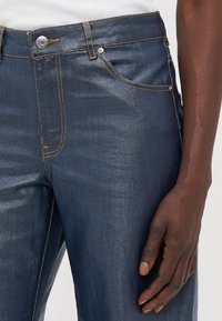 Donkerblauwe spijkerbroek van denim met een gestructureerde afwerking, voorzien van gouden stiksels, een zilveren knoop en een voorkantzak. Hand rustend op de tailleband.