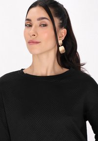 Top nero testurizzato con un motivo geometrico, dotato di un collo ampio. La modella indossa grandi orecchini dorati con una finitura opaca.