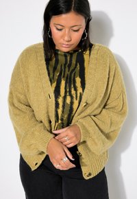 Khaki oversize pletený kardigan s žebrovanými manžetami a dřevěnými knoflíky, vrstvený přes černý a žlutý top s tygřím vzorem. Viditelné černé džíny.