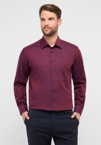 Camicia bordeaux a maniche lunghe con colletto appuntito, taschino frontale e chiusura con bottoni; tessuto liscio con una leggera lucentezza.