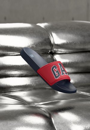 Sandalo a infradito rosso e blu navy con logo "GAP" che fluttua sopra un divano argentato, metallico e trapuntato.