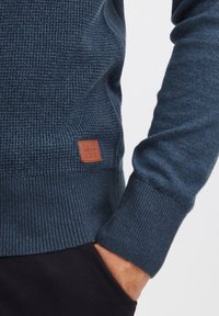 Pullover di lana blu scuro con polsini e vita a costine. Presenta un'etichetta in pelle marrone sul lato. Texture liscia e design aderente.