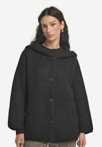 Mörkgrå huva cardigan gjord av texturerad stickad tyg, med en framknäppning, sidofickor och vida ärmar.