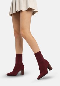 Botines de punto burdeos hasta el tobillo con punta afilada y ajuste tipo calcetín, con un tacón grueso de madera y parte superior acanalada y texturizada.