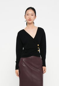 Sweater preto envolvente com decote em V, apresentando três botões dourados na cintura. Combinando com uma saia de couro brilhante em bordeaux. Textura suave.