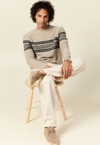 Suéter de punto beige con patrón geométrico negro, pantalones de un beige claro y zapatos de ante color beige. Modelo sentado en un taburete de madera, en una pose relajada.