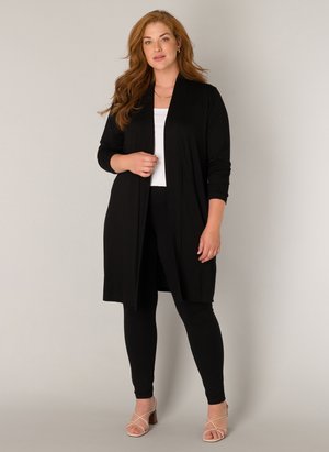 Frau mit langem roten Haar, die eine schwarze offene Strickjacke, ein weißes Oberteil, schwarze Leggings und beige Sandalen mit Absatz trägt, vor einem einfarbigen Hintergrund stehend.