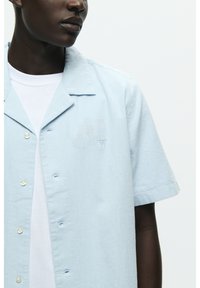 Chemise à manches courtes bleu clair en tissu lisse avec un col camp, dotée de boutons blancs et d'un petit logo brodé sur la poitrine.