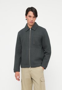 Grå zip-up jakke med kragehals, med en brystlomme og solid tekstur. Brukt med beige cargo-bukser.