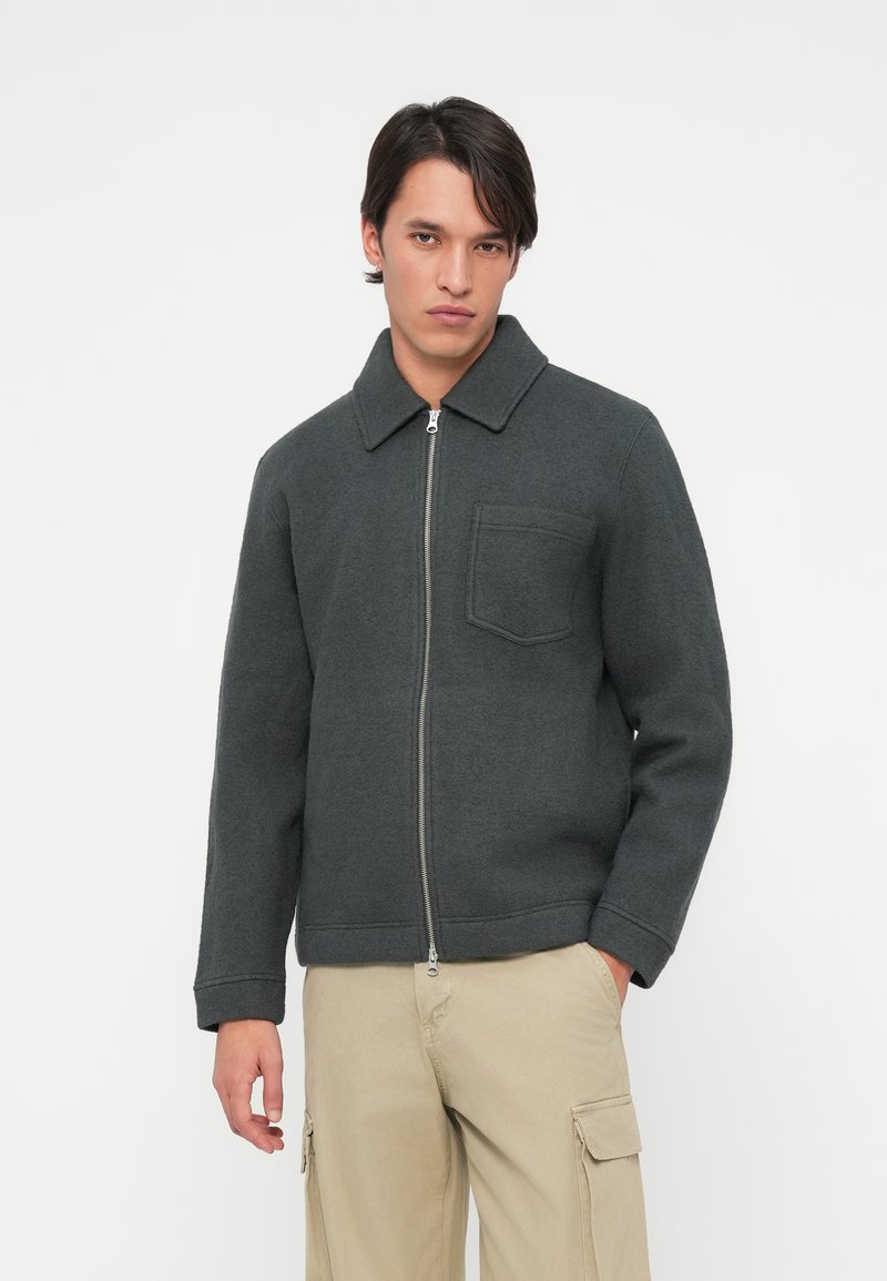 Grå zip-up jakke med kragehals, med en brystlomme og solid tekstur. Brukt med beige cargo-bukser.