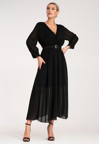 Czarna sukienka z głębokim dekoltem w kształcie V, długimi przezroczystymi rękawami i paskiem w talii. Wykonana z lekkiego materiału z warstwową spódnicą. Czarne sandały na obcasie.