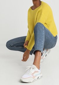 Osoba oblečená v žlutém svetru, modrých džínách a bílých teniskách, která se krčí proti jednobarevnému světlému pozadí.