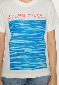 T-shirt de algodão com um corpo branco, apresentando uma impressão de água azul e texto vermelho no topo: "OS MARES VÃO SUBIR E NÓS VAMOS TODOS NOS AFUNDAR."