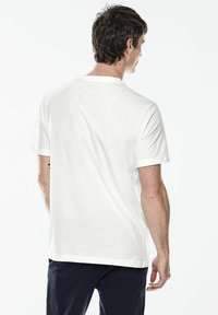 Bílý tričko s kulatým výstřihem, krátké rukávy, měkký materiál, volný střih, jednoduchý design, bez viditelných vzorů, viditelné švy, nošené s tmavými kalhotami.