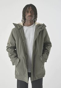 Olivgrüner Parka mit einem weichen, beigen Fleece-Futter, Fronttaschen und einem Reißverschluss. Mit Kapuze und Druckknöpfen gestaltet.