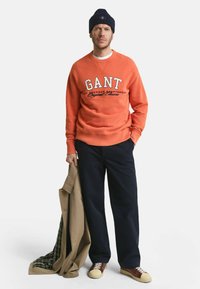 Oranje sweatshirt met "GANT" logo, donkerblauwe broek, beige jas met geruite voering en sneakers met crèmekleurige accenten.