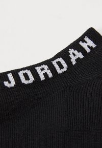 Calcetín negro con textura acanalada, que presenta "JORDAN" en blanco tejido en el puño. Diseño simple sin patrones ni acentos adicionales.