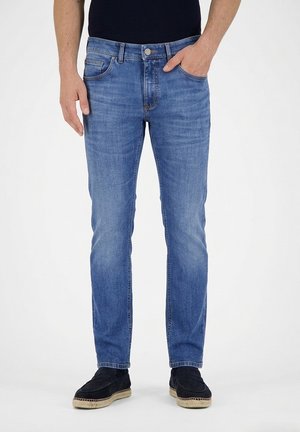 Man wearing blue denim jeans and black slip-on shoes, standing with one hand in pocket against a plain white background.