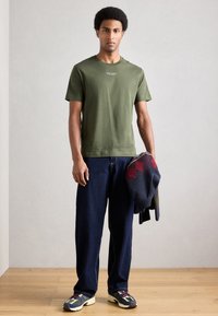 Χακί βαμβακερό μπλουζάκι, άνετο σκούρο μπλε τζιν παντελόνι και πολύχρωμα αθλητικά παπούτσια. Κρατάει ένα μαύρο και κόκκινο πουλόβερ με σχέδια.