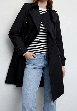 Osoba w czarnym dwurzędowym płaszczu przeciwdeszczowym, czarno-białej koszulce w paski i jasnoniebieskich dżinsach, z jedną ręką w kieszeni, stoi przy białej ścianie.