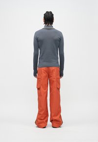 Garām piedurknēm pelēks tops un plaša griezuma oranžas kravas bikses. Bikšu sānos ir kabatas, kontrastējošas svītras un gluda tekstūra.