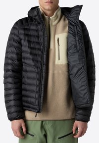 Abrigo acolchado negro con diseño acolchado y capucha, sobre un pulóver de forro polar beige con un bolsillo con cremallera, combinado con pantalones verdes.