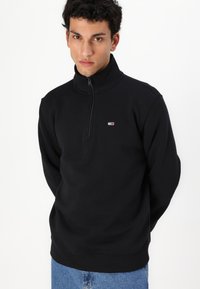 Zwarte ritssluiting sweatshirt gemaakt van zacht materiaal, met een hoge kraag en een klein logo op de borst. Gecombineerd met blauwe jeans.