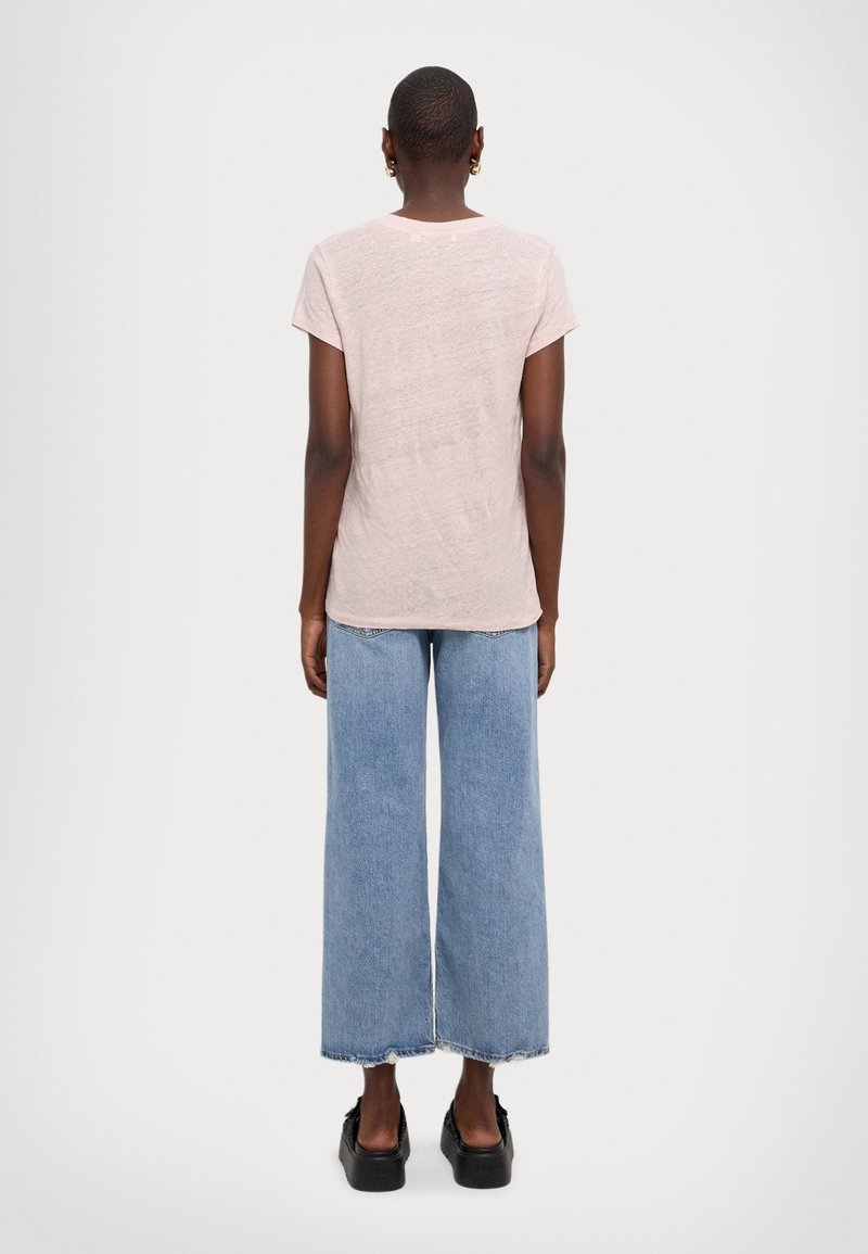 Osoba z krótkimi włosami ubrana w jasnoróżową koszulkę z krótkim rękawem, szerokie niebieskie dżinsy, czarne buty na platformie i złote kolczyki w kształcie kół, stojąca tyłem.