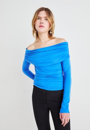 Mėlyna off-the-shoulder viršutinė dalis, pagaminta iš tempimo medžiagos, su susiraukšlėjimo detalėmis per krūtinę ir ilgomis rankovėmis. Suderinta su juodomis kelnių dalimis.