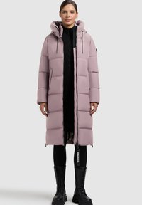 Lang lyserød pufferjakke med stor hætte, kviltet design, front lynlås og sidelommer, parret med sort ribbestrikket sweater og knæhøje støvler.