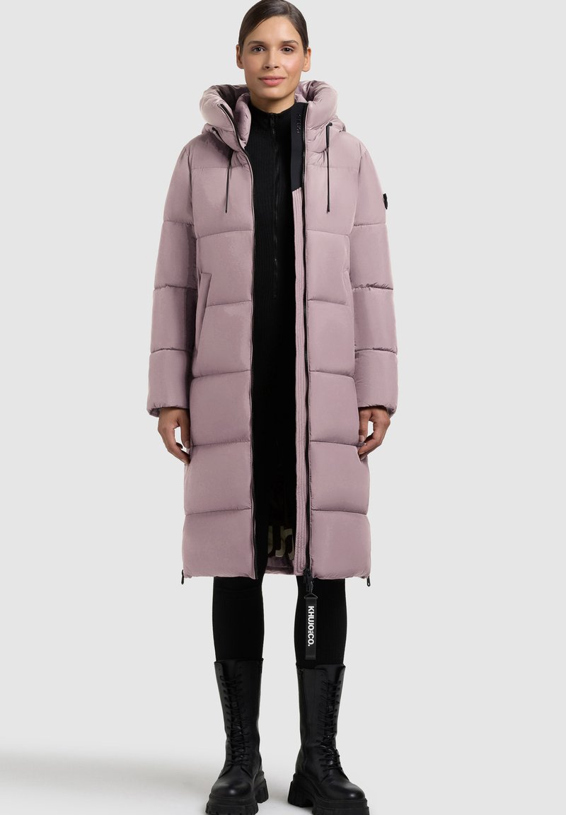 Lang lyserød pufferjakke med stor hætte, kviltet design, front lynlås og sidelommer, parret med sort ribbestrikket sweater og knæhøje støvler.