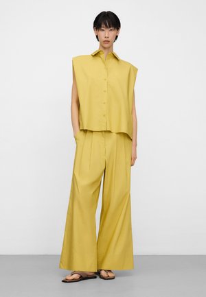 Osoba ubrana w bezrękawnik w kolorze musztardowym z guzikami oraz szerokie spodnie w tym samym kolorze, ubrana w brązowe sandały, stojąca na jasnym, jednolitym tle.