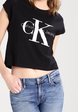 Schwarzes, kurzes T-Shirt mit kurzen Ärmeln, das ein großes weißes "CK"-Logo und den Text "Calvin Klein Jeans" aufweist; getragen mit hellblauen Jeans.