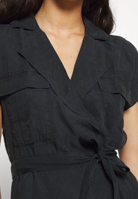 Abito avvolgente nero con colletto, maniche corte, due tasche sul petto e vita legata. Tessuto morbido e liscio.