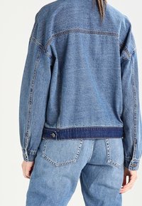 Denim jacket in light blue with a loose fit, featuring a dark blue band at the waist and visible stitching on the back.